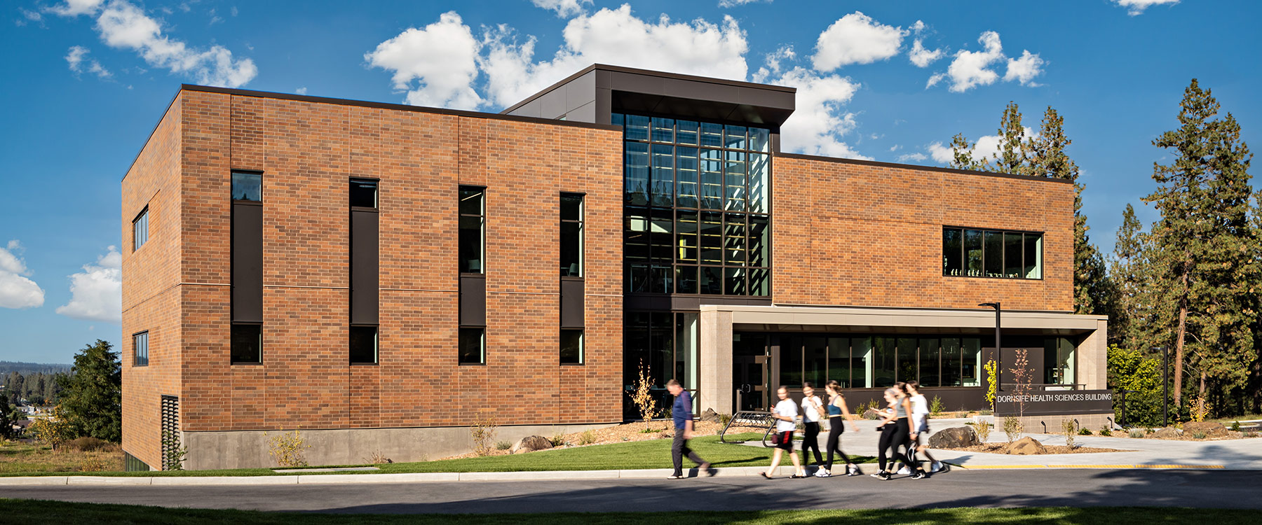 The outside of a 38,000-square-foot, state-of-the-art health sciences building. The building is modern-looking with large windows, surrounded by green grass. Students stand outside, conversing with each other.